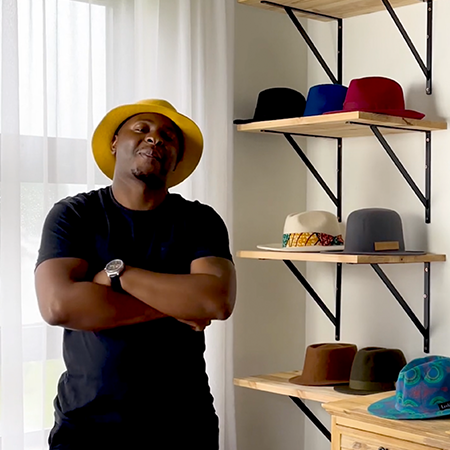 MediaCom's Brian Muguto wearing a fedora next to a wall of similar hats